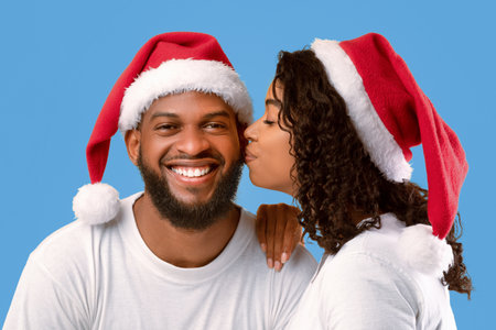 Loving african american woman in Santa hat kissing her smiling husband, posing over yellow studio backgroundの写真素材