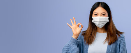 Woman Wearing a Mask Shows Okay Gesture With Hand Against a Blue Backgroundの写真素材