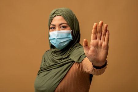 A Young Muslim Lady Wearing a Protective Mask and Hijab Stands Against a Brown Backgroundの写真素材