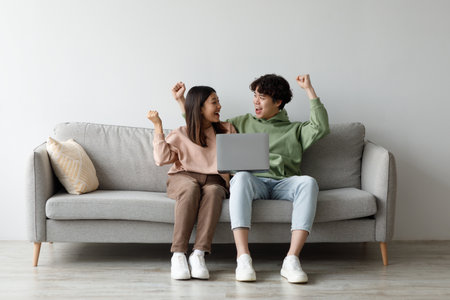 Young Asian Couple Celebrates Success Together at Home With Laptop on Sofaの写真素材