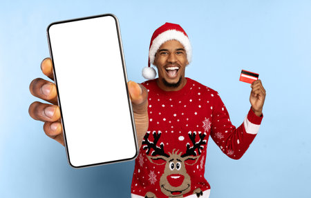 Excited Man in Christmas Sweater Shows Smartphone and Credit Card for Holiday Shopping