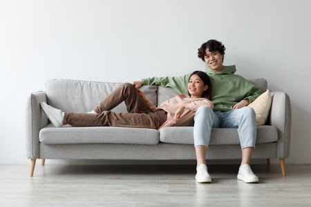 Cheerful Millennial Asian Couple Enjoying Cozy Weekend on Couch, Relaxing Together at Homeの写真素材