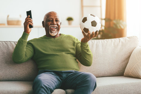 Senior Man Enjoying Soccer Match at Home While Holding Remote and Ballの写真素材