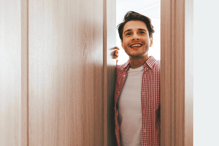 Young Man Smiling While Opening a Wooden Door in a Bright Roomの写真素材