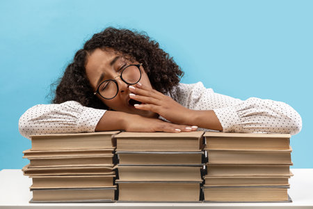 Young African American Woman Yawning and Feeling Bored While Studying for Exam on Blue Backgroundの写真素材