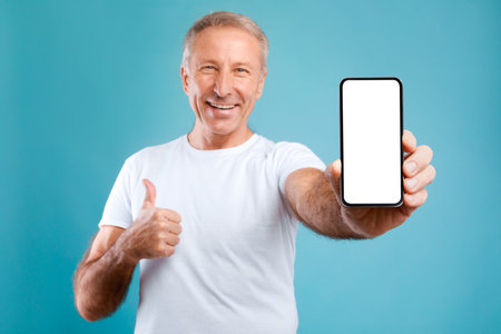 Mature Man Happily Holds Blank Smartphone Screen and Gives Thumbs up in Bright Blue Settingの写真素材