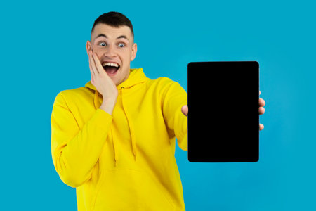 Young Man in Yellow Hoodie Reacts Excitedly While Holding a Tablet Against a Bright Blue Background in a Joyful Moment of Discoveryの写真素材