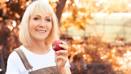 Senior Woman Holding a Red Apple Outdoors in a Sunny Autumn Gardenの写真素材