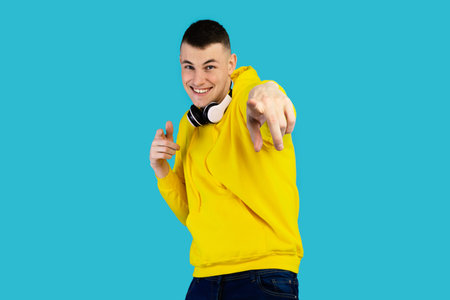 Young Man With Headphones Wearing Yellow Hoodie Poses Playfully Against Bright Blue Backgroundの写真素材
