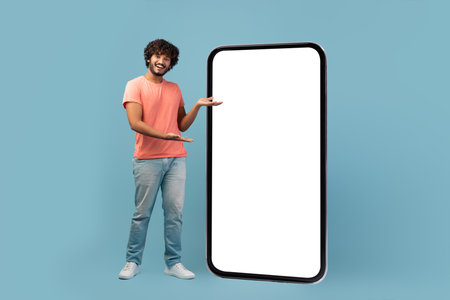Stylish Young Eastern Man Standing Beside Large Blank Cell Phone Mockup in Blue Backgroundの写真素材