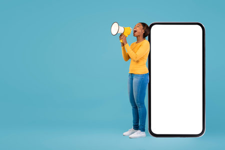 Young Woman Using a Megaphone Beside a Large Blank Smartphone on a Blue Background in a Vibrant Setting for Communication Cultureの写真素材