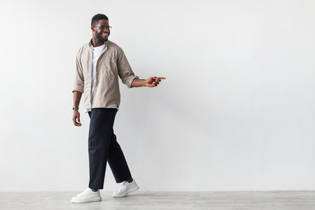 Happy Young Black Man Walking Against a White Wall, Pointing to Free Space for Promotional Textの写真素材