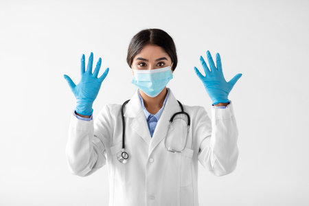 Calm Young Lady Doctor in Coat Shows Gloved Hands While Wearing a Protective Mask Indoorsの写真素材