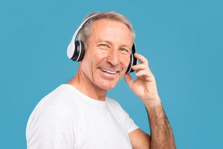Mature Man Enjoys Music Wearing Wireless Headphones Against a Blue Backgroundの写真素材