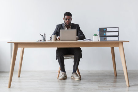 Focused Young Black Man Working Remotely at a Desk With Laptop Against White Wallの写真素材