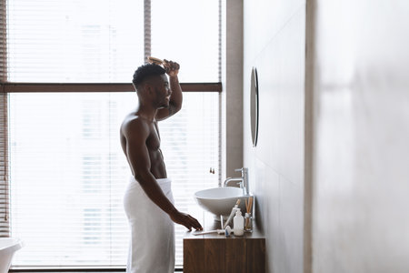 African Guy Brushes Hair Near Mirror in Modern Bathroom During Morning Routineの写真素材