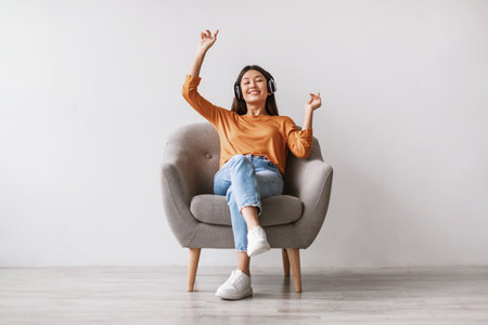 Young Asian Woman Enjoys Music While Dancing in Armchair Against White Studio Wallの写真素材