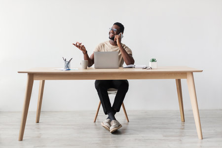 Young Black Man Multitasking in Home Office, Using Laptop and Cellphone for Remote Communicationの写真素材