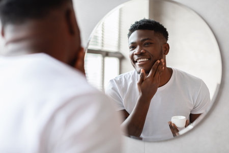 Smiling African Man Applying Moisturizer in Bathroom While Looking at Reflection in Mirrorの写真素材