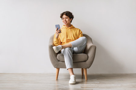 Young Asian Man Enjoying Social Media on His Cellphone While Relaxing in an Armchairの写真素材