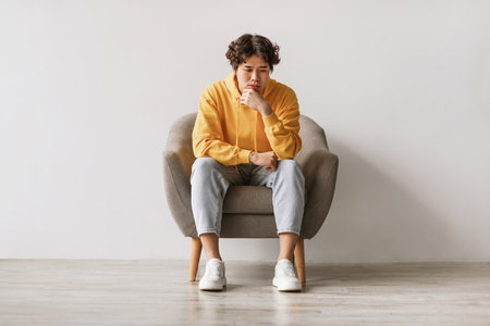 Thoughtful Young Asian Man Sitting in Armchair Feeling Sad and Overwhelmed With Stressの写真素材
