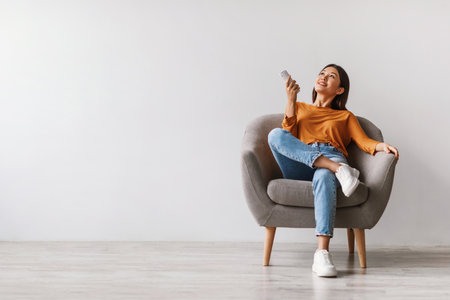 Young Woman Relaxes With Remote in Armchair Under Air Conditionerの写真素材