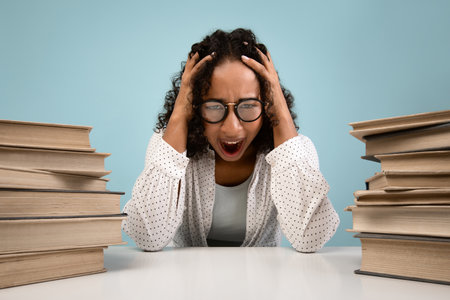 Stressed Student Cries out in Frustration Over Piles of Books in Blue Studio Backgroundの写真素材
