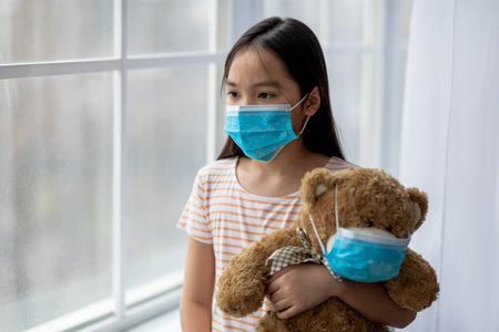 Child Holding Teddy Bear While Looking out Window Wearing Face Mask During Pandemicの写真素材