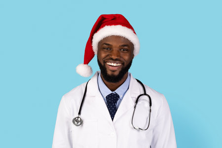 Handsome black doctor in uniform and Santa hat posing over blue backgroundの写真素材