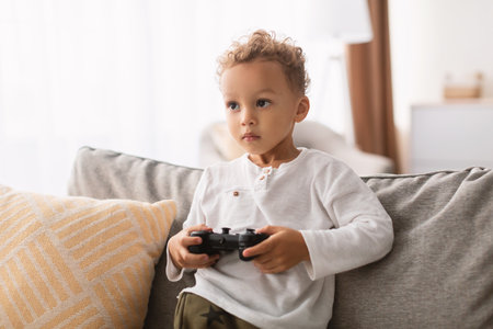 African American Boy Enjoys Playing Video Game Indoors With Excitement on the Sofaの写真素材