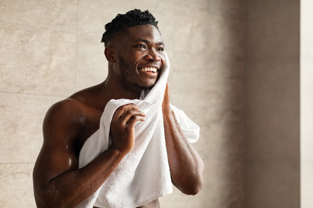 Handsome Black Man Enjoying Morning Skincare Routine in Modern Bathroom While Wiping Faceの写真素材