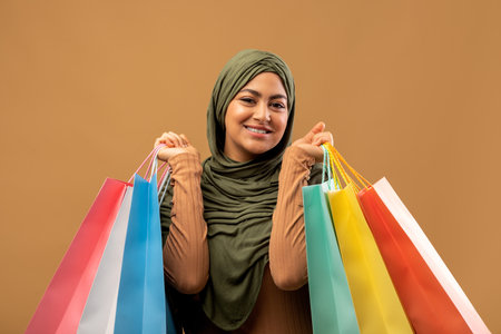 Seasonal sales. Happy arab woman in hijab holding lots of shopping bags, standing over brown studio backgroundの写真素材