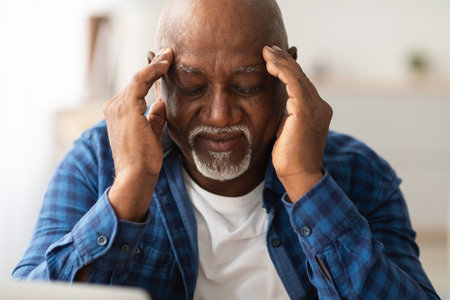 Senior African American Man Massages Temples While Dealing With Headache Indoors During Daytimeの写真素材