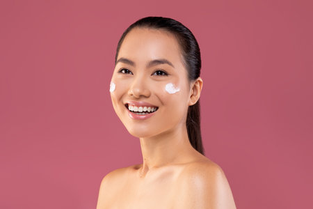 Happy Young Woman Testing Moisturizer Cream on Her Face in a Vibrant Studio Settingの写真素材