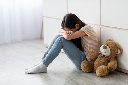 Young Girl Sitting on the Floor With Her Teddy Bear in a Room, Feeling Sad and Isolated During an Emotional Momentの写真素材