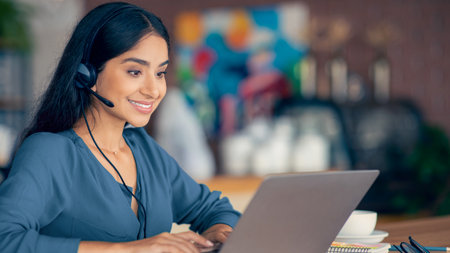Woman Working on Laptop With Headset in Busy Cafe Environmentの写真素材