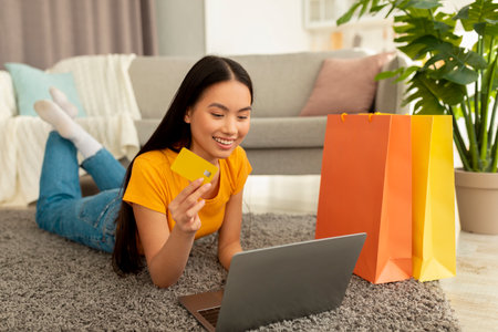 Happy Korean Woman Shopping Online With Laptop and Bags in Cozy Home Settingの写真素材