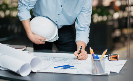 Construction Professional Drafting Plans at a Worksite in an Office Setting While Holding a Hard Hatの写真素材