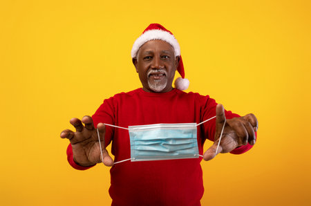 Happy Senior Black Man Wearing a Santa Hat Joyfully Holds a Medical Face Mask in Outstretched Handsの写真素材