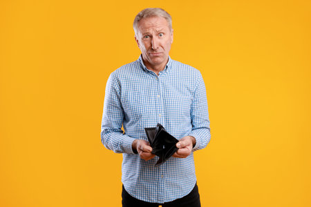 Sad Older Man Stands Alone in Front of an Orange-Yellow Background While Holding His Empty Wallet