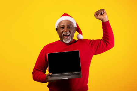 Senior Man in a Santa Hat Celebrates Online Christmas Sale With a Laptop on an Orange Backdropの写真素材