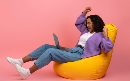 Celebrating Success, Young Black Woman Raises Arms in Joy While Using Laptop on Pink Backdropの写真素材