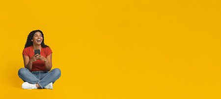 Happy Woman Sitting on Floor Using Smartphone Against Yellow Background
