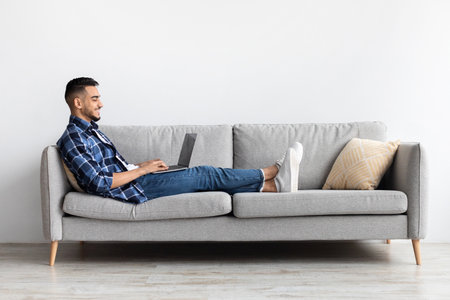 Smiling Arab Man Enjoys Using Laptop on Couch at Home for Leisure and Online Activitiesの写真素材