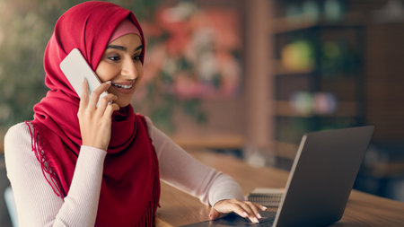 Young Woman in a Red Hijab Talks on the Phone While Working on a Laptop in a Cozy Setting