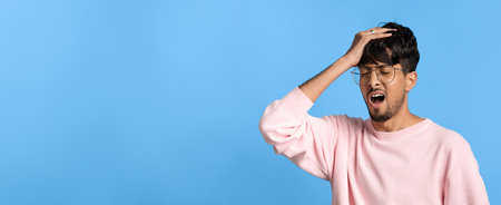 Young Man Expressing Frustration Against a Blue Background While Wearing Glasses and a Pink Sweaterの写真素材