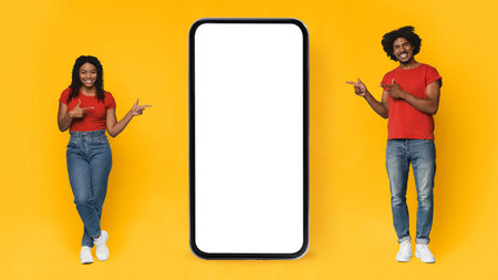 Happy Young Couple Pointing at a Blank Smartphone Against a Vibrant Yellow Backgroundの写真素材