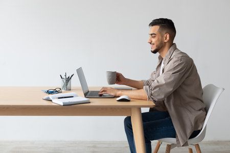 Smiling Arab Man at Home Using Laptop and Enjoying Coffee While Working at Deskの写真素材