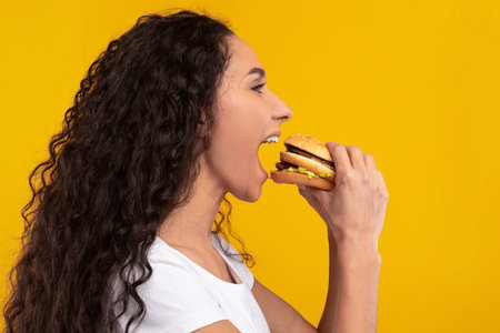 Funny Hungry Lady Biting Into a Big Burger Against a Bright Yellow Backgroundの写真素材