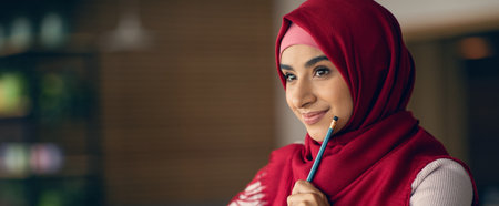 Young Woman in Red Hijab Holds Pencil and Smiles While Contemplating Her Ideas in a Cozy Settingの写真素材